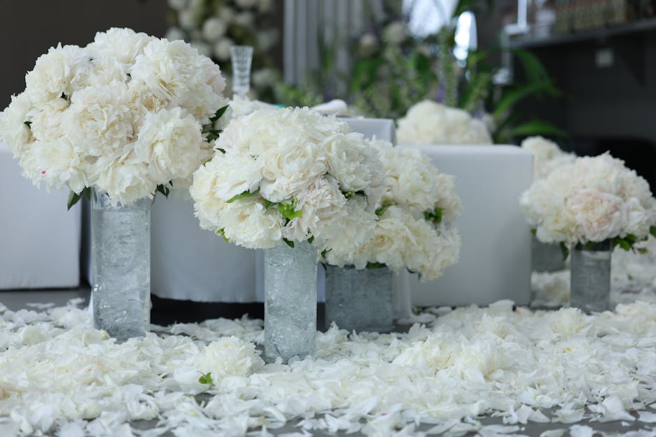 A series of elegant floral arrangements featuring large, densely packed white peonies and hydrangeas, displayed in tall, clear glass vases with a textured surface. The vases are placed on a surface co