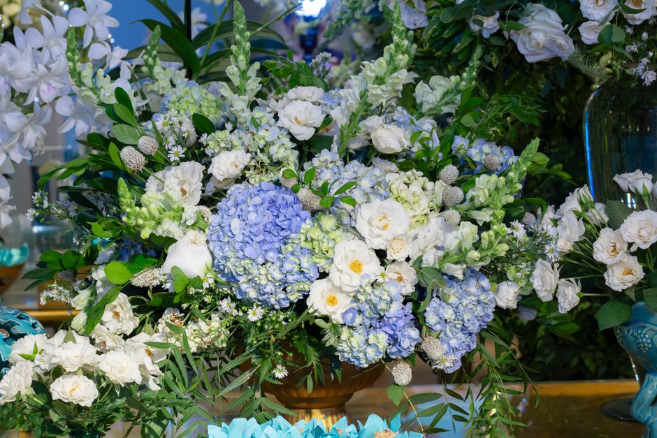A large floral arrangement featuring predominantly white and pale blue flowers, including hydrangeas, roses, and small daisy-like blooms, arranged with lush green foliage. The flowers are arranged in 