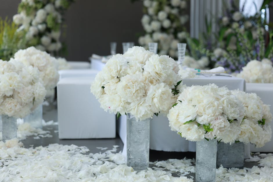 A floral arrangement featuring large, rounded bouquets of white peonies and hydrangeas placed in tall, rectangular glass vases on a table covered with white petals and flower confetti. The flowers app