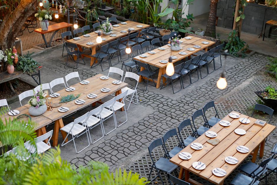 An outdoor dining area featuring three long wooden tables set for a meal, surrounded by black and white chairs. Each table is decorated with small floral arrangements in round, textured grey vases tha
