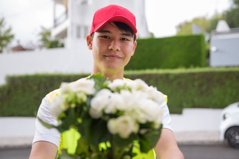 A young man wearing a red cap and a high-visibility vest is outdoors, holding a bouquet of fresh white roses and tulips wrapped in clear paper with green foliage. He is smiling gently and offering the