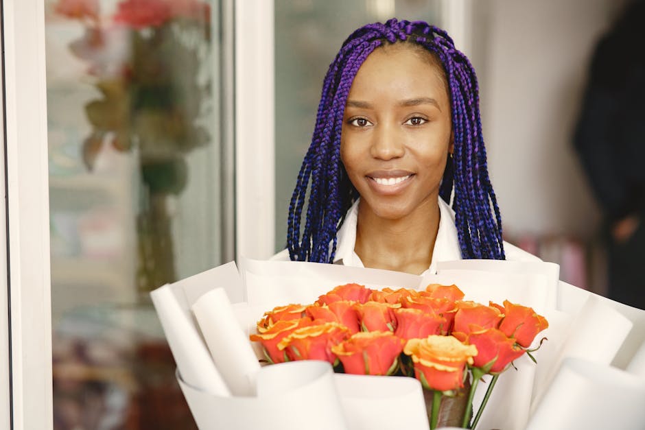 A smiling woman with vibrant purple braids holds a large bouquet of fresh roses, predominantly in shades of orange and coral, wrapped in white floral paper. She is standing indoors near a window, with