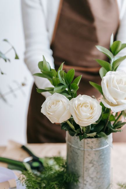 A florist arranging a bouquet of fresh white roses with lush green foliage, set in a metallic silver vase on a wooden surface. The background includes blurred elements of the workspace, with natural l